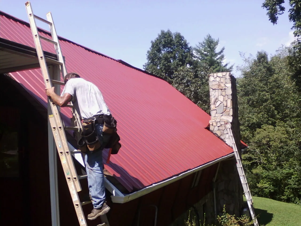 Roofer on a ladder beside a red metal roof and stone chimney — Soffit & Fascia Repair by Roofing Brothers in Rockland